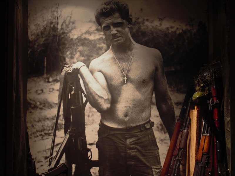 A black and white photograph of shirtless man with cross necklace holding firearms.